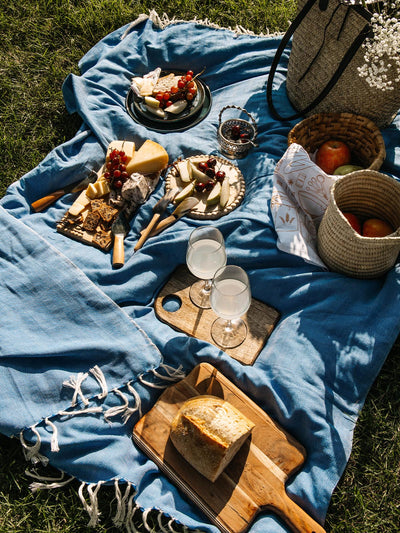 Outdoor picnic setup on a blue blanket with a variety of cheeses, bread, fruits, and wine, laid out on a wooden board and plates, with woven baskets and a bag in the background