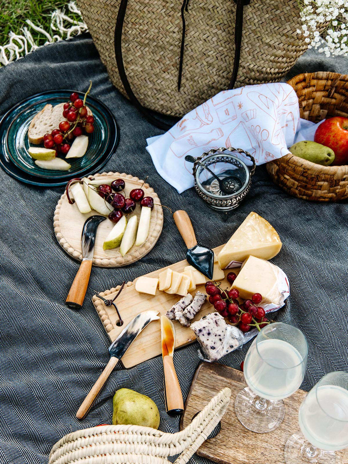 Close-up of an outdoor picnic spread on a dark gray blanket, featuring cheese, fruits, and bread on wooden boards, surrounded by a woven basket, bag, and picnic accessories.