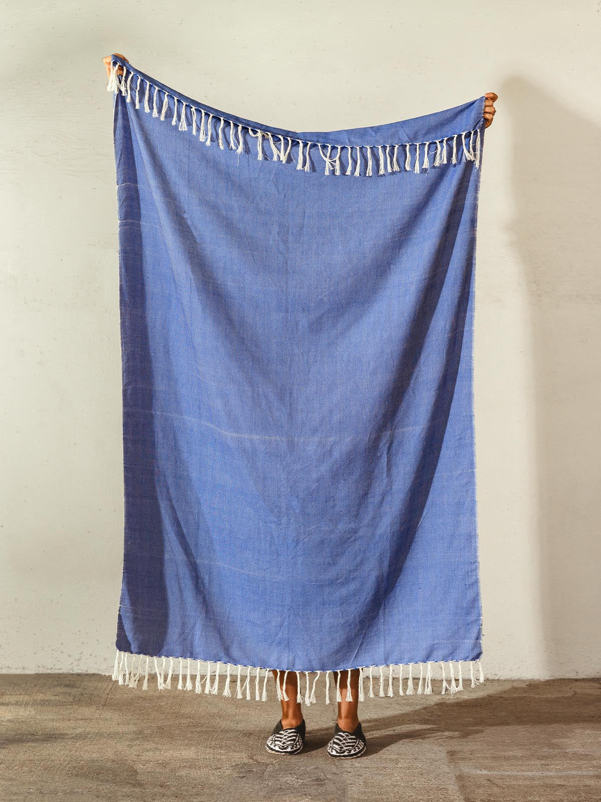 Handwoven blue cotton blanket with white fringe held up by a person, showcasing the full size and intricate craftsmanship.