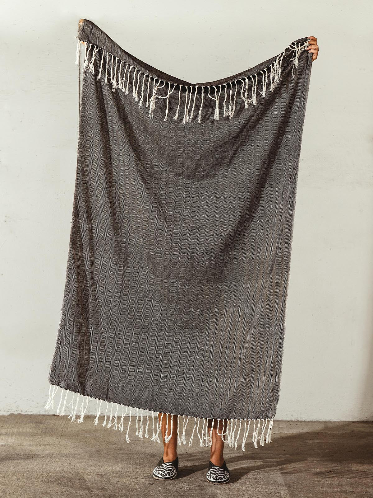 Handwoven dark gray cotton blanket with white fringe displayed by a person, highlighting the texture and design.