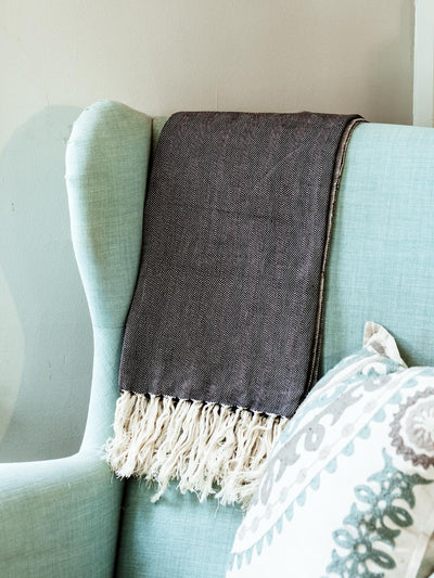 Gray herringbone pattern cotton blanket with white fringes draped over the back of a light teal upholstered armchair, with a decorative pillow in the foreground.