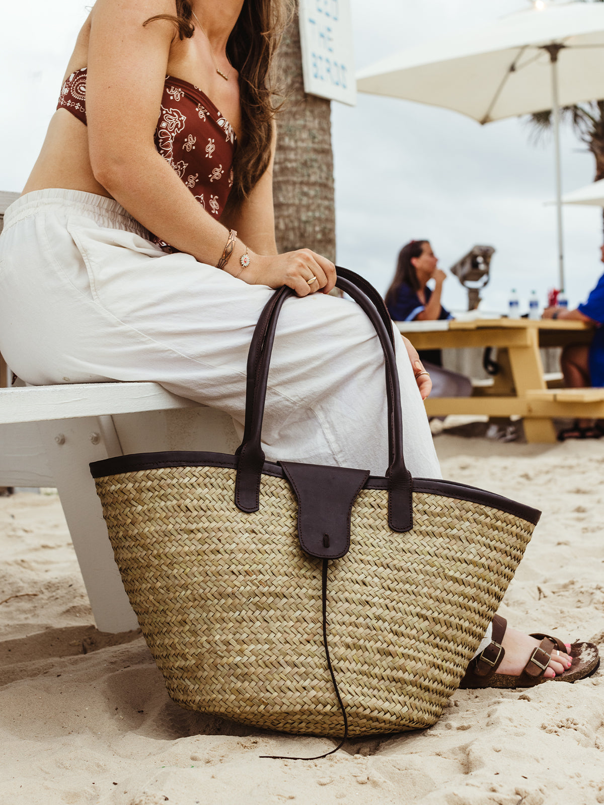 Close-up of a large straw tote bag with black leather handles, resting on a woman's lap as she sits on a beach bench, dressed in a maroon bandeau top and white wide-leg pants.