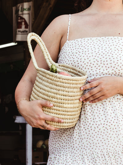 Close-up of a woman in a white floral sundress, holding a small woven basket filled with fresh produce, with a focus on her gold rings and bracelets.