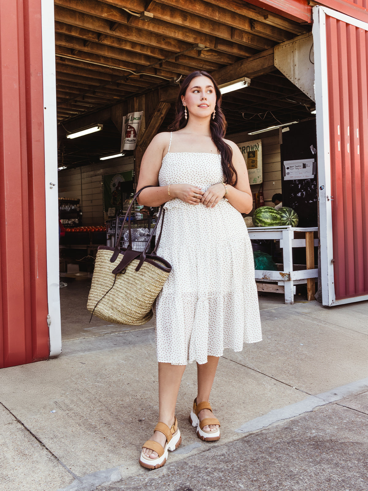 Woman in a white floral sundress, standing in front of a market with a large straw tote bag, wearing tan platform sandals and hoop earrings.