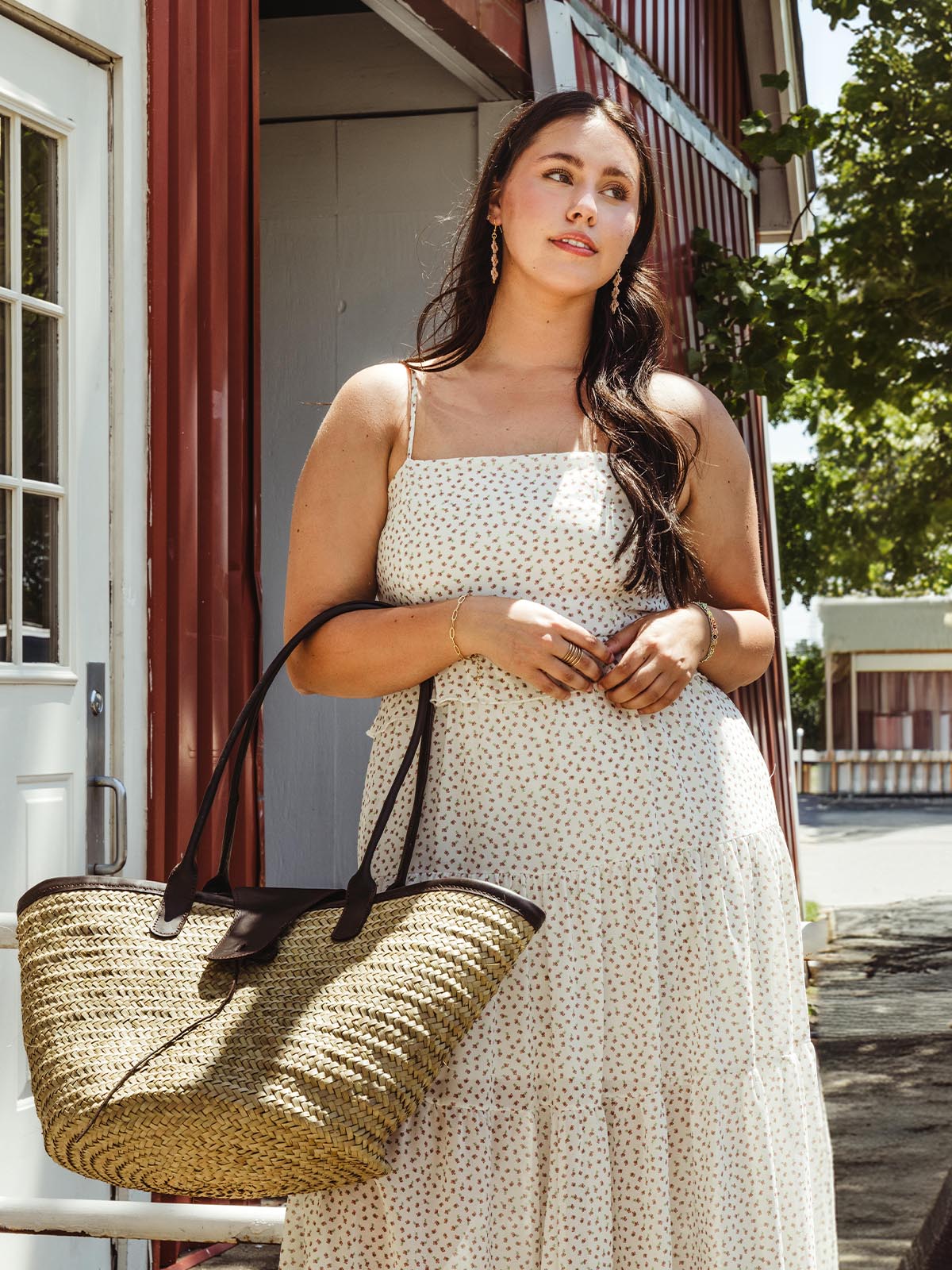 Woman wearing a white sundress with a small floral pattern, holding a large straw tote bag with black leather handles, standing outside a red barn-style building.