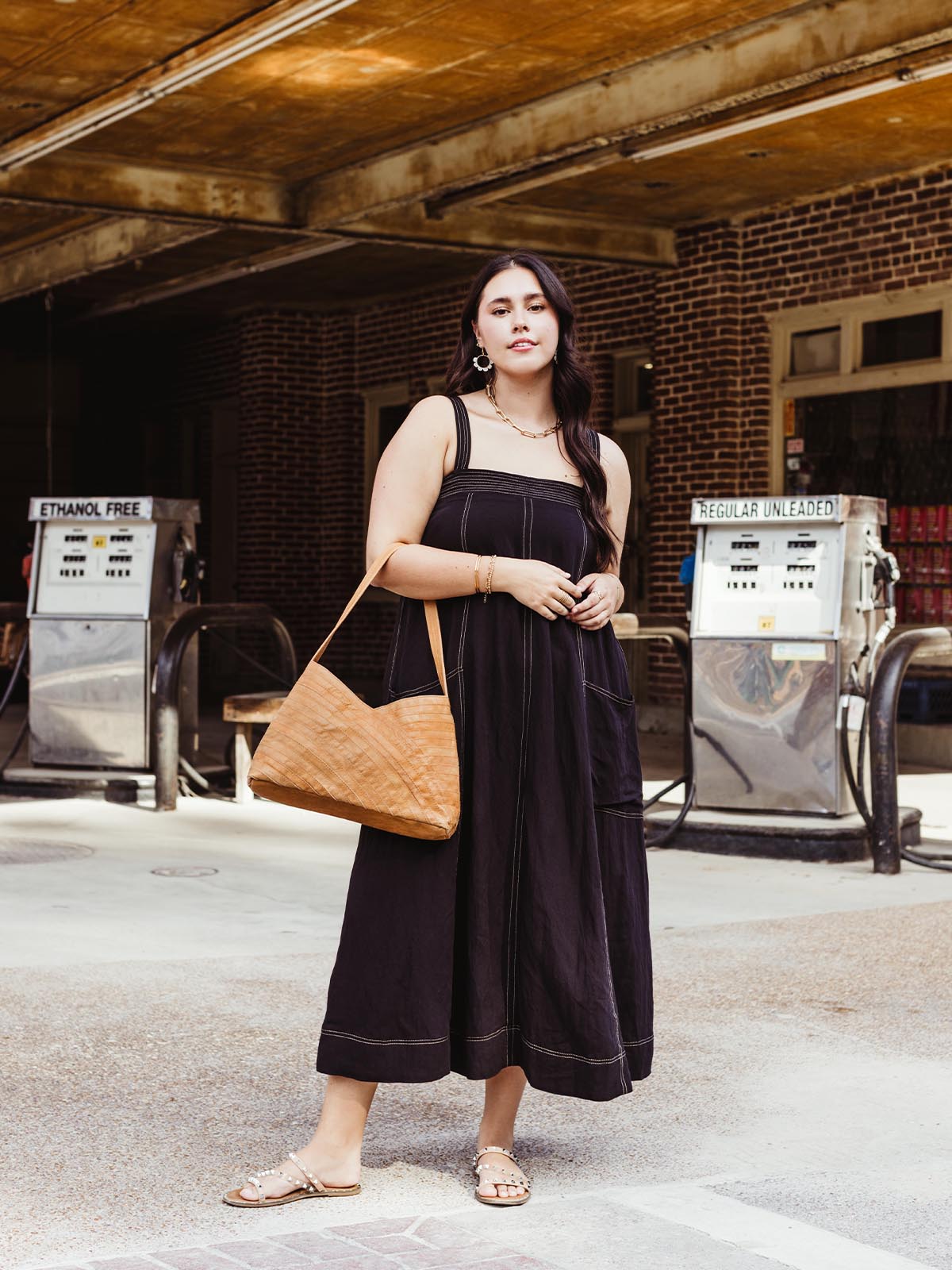 Woman dressed in a black sundress, standing in front of vintage gas pumps, carrying a tan leather shoulder bag with a chevron pattern, wearing pearl-studded sandals and gold jewelry.