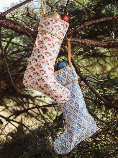 Pair of handcrafted Christmas stockings, one with a red leaf pattern and the other with a blue leaf pattern, hanging together on a pine tree branch.