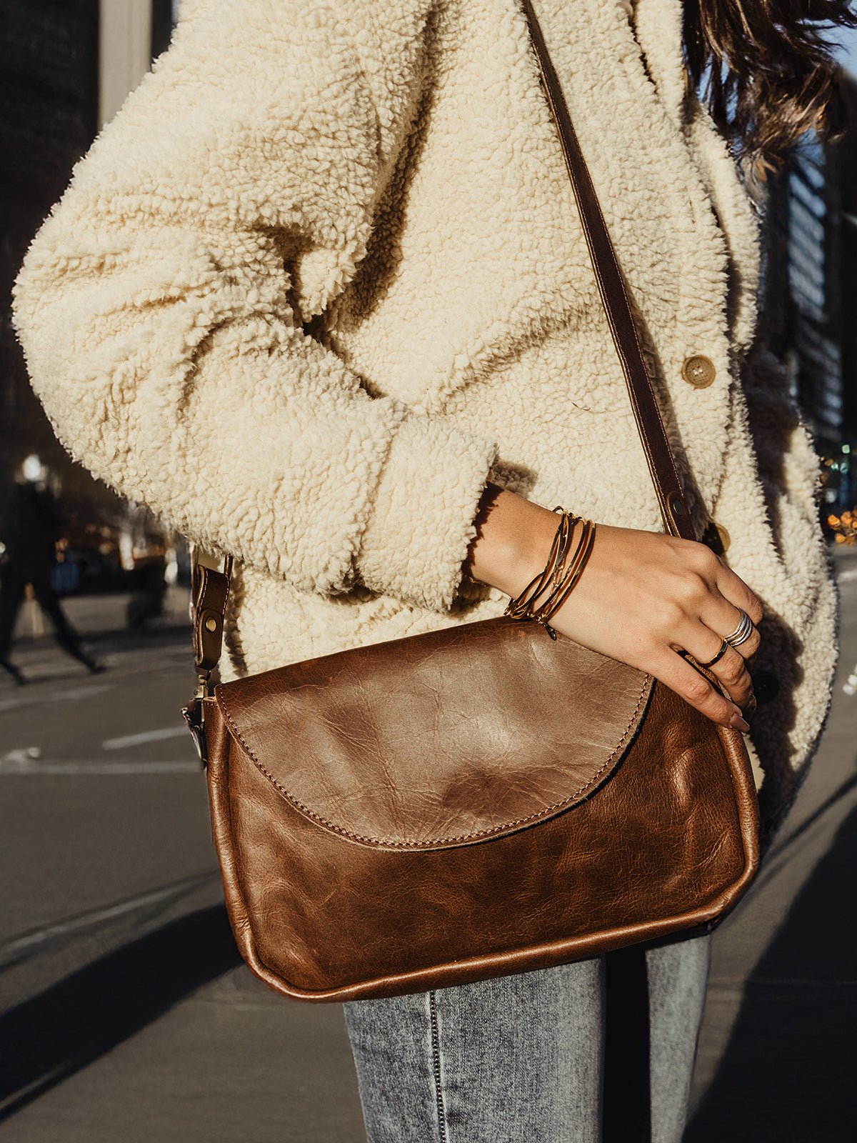 A close-up of a woman dressed in a cozy cream sherpa jacket, carrying a compact brown leather crossbody bag with a curved flap design. She accessorizes with minimal jewelry, including bracelets and rings, while the urban street and soft evening light create a warm atmosphere.