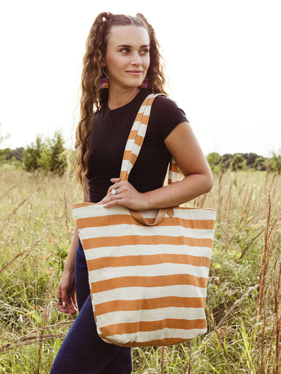 Female model in an outdoor setting  holding orange and cream striped canvas tote. 