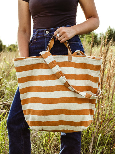 Female model in a outdoor setting  holding orange and cream striped canvas tote. 