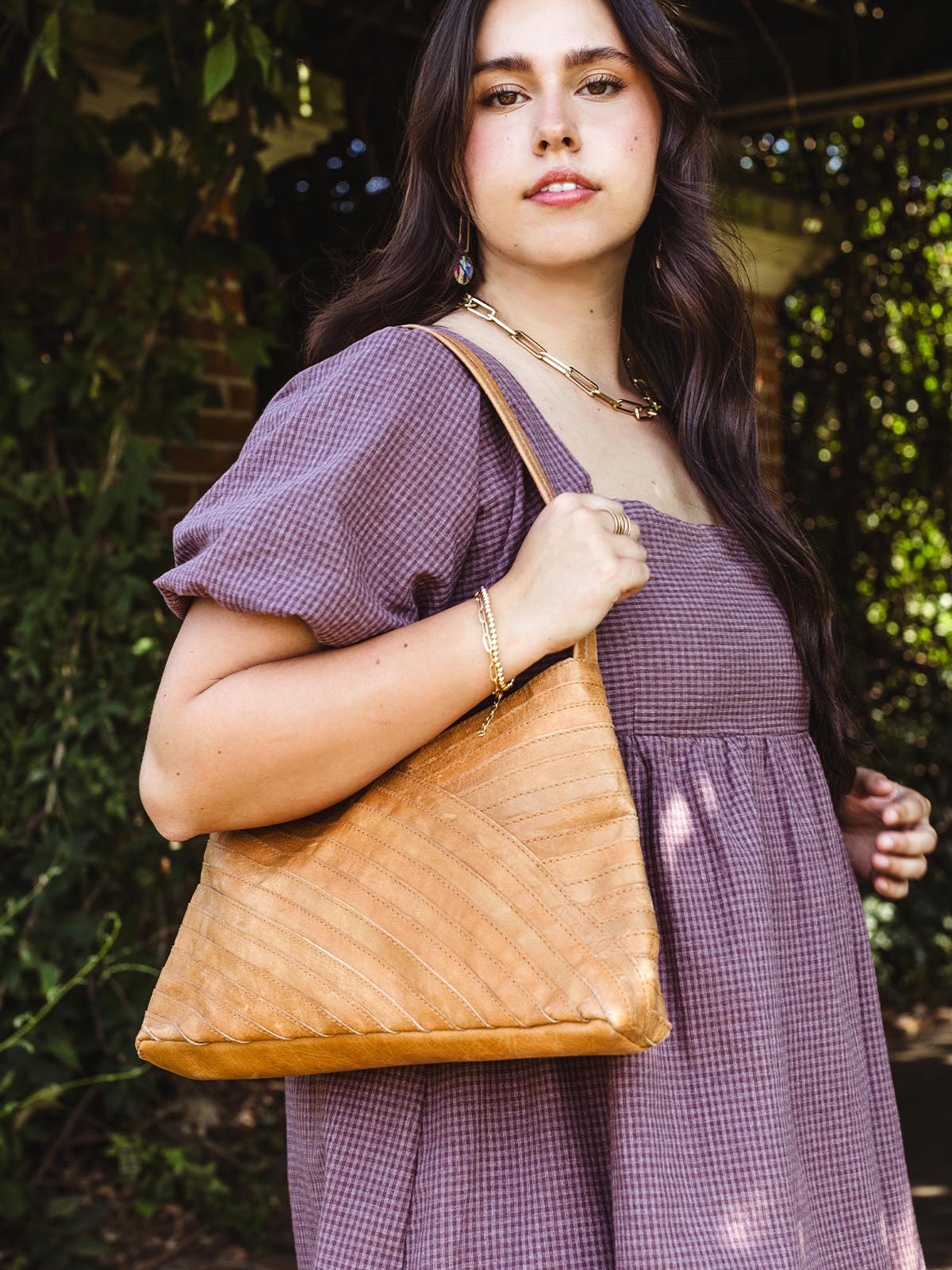 Woman wearing a mauve puff-sleeve dress, carrying a tan leather shoulder bag with a chevron pattern, accessorized with a gold chain necklace and earrings, standing in front of lush greenery.