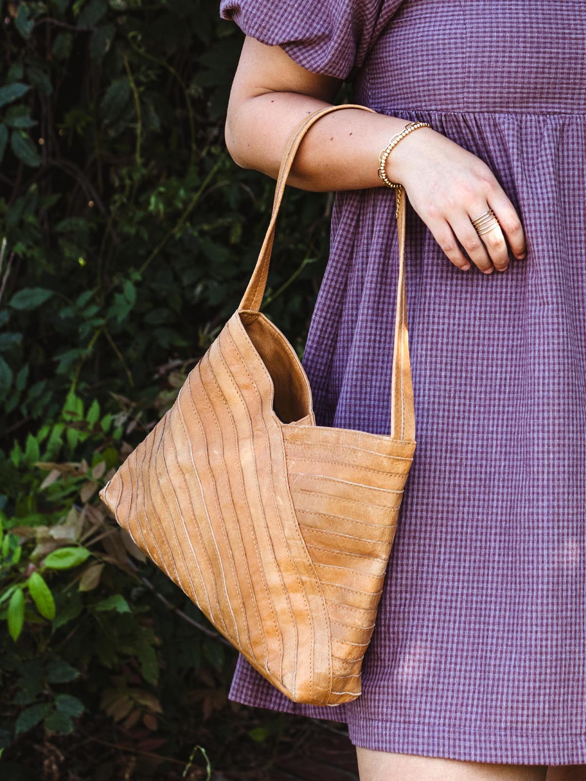 Close-up of a woman in a mauve puff-sleeve dress, holding a tan leather shoulder bag with a chevron pattern, highlighting her gold bracelet and rings.