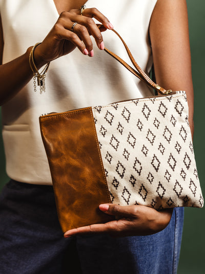 Woman holding a stylish leather and canvas clutch bag with a geometric black and white pattern, featuring a brown leather accent and wrist strap. Perfect for everyday elegance.