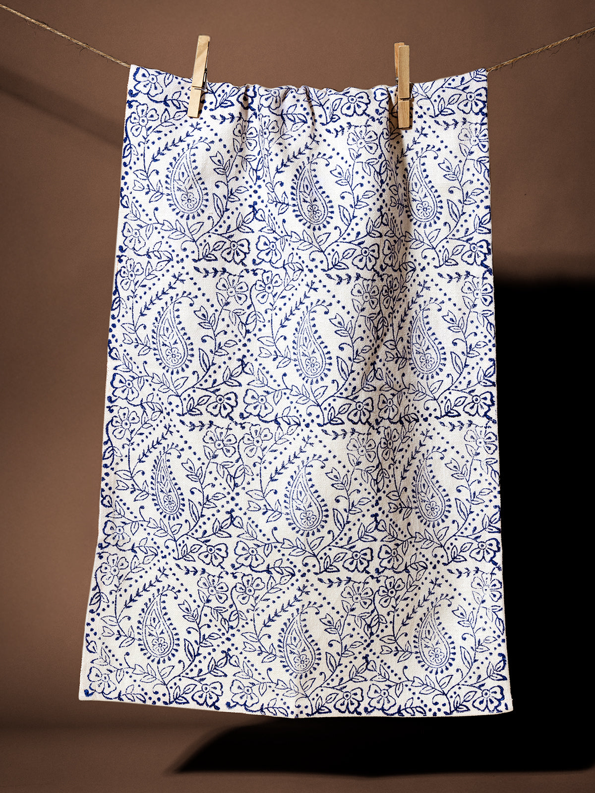 Handcrafted blue paisley tea towel hanging on a clothesline against a brown background. Ethically made with intricate floral and paisley patterns, perfect for kitchen decor or gifting.