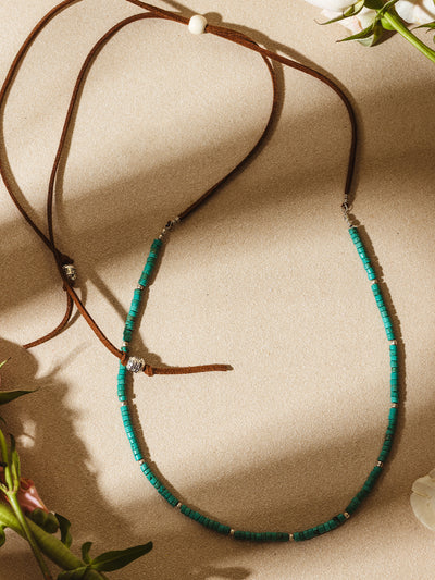 Flat lay of a green beaded necklace with silver accents and brown suede cord, styled with soft shadows and floral accents on a beige background.