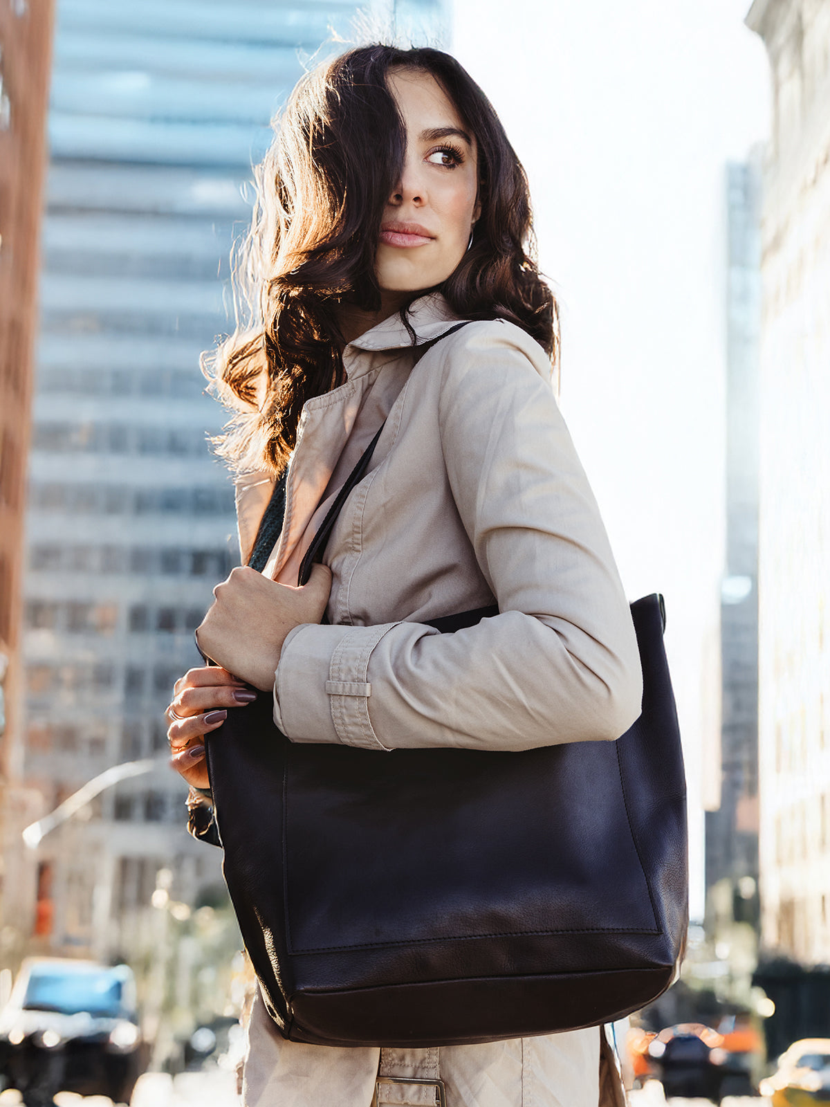 A stylish woman stands outdoors in a sunlit urban setting, wearing a light beige trench coat and carrying a sleek black leather tote bag on her shoulder. The cityscape, with tall buildings and sunlight filtering through, serves as the backdrop.