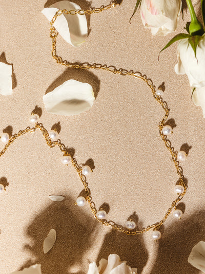 Flat lay of a gold chain necklace adorned with dangling white freshwater pearls, styled among white rose petals on a softly lit neutral background.