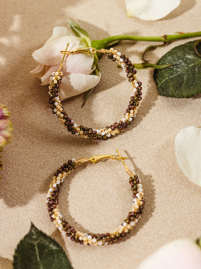 Flat lay of handcrafted hoop earrings made with gold wire and warm-toned beads, styled beside a pale pink rose and scattered flower petals on a soft neutral surface.
