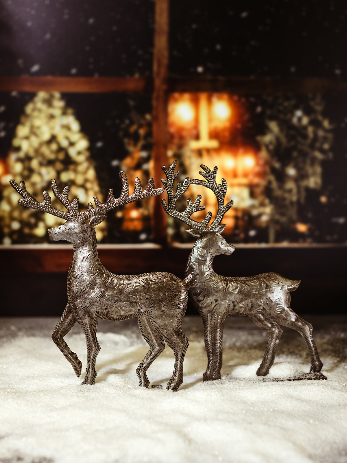 Two handmade metal reindeer statues standing on a snow-covered surface, with a glowing holiday background, showcasing intricate detailing on the antlers and bodies.