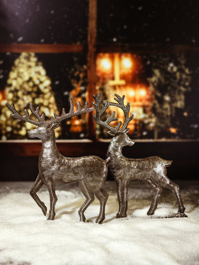 Two handmade metal reindeer statues standing on a snow-covered surface, with a glowing holiday background, showcasing intricate detailing on the antlers and bodies.