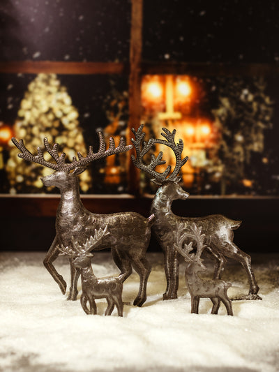 Set of four handcrafted metal reindeer statues in various sizes, arranged on a snow-covered surface with a warm holiday backdrop, highlighting their detailed antlers and rustic finish.