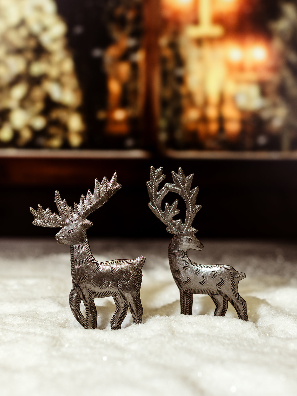 Two small handcrafted metal reindeer statues with intricately designed antlers and bodies, standing on a snow-covered surface with a cozy holiday background.