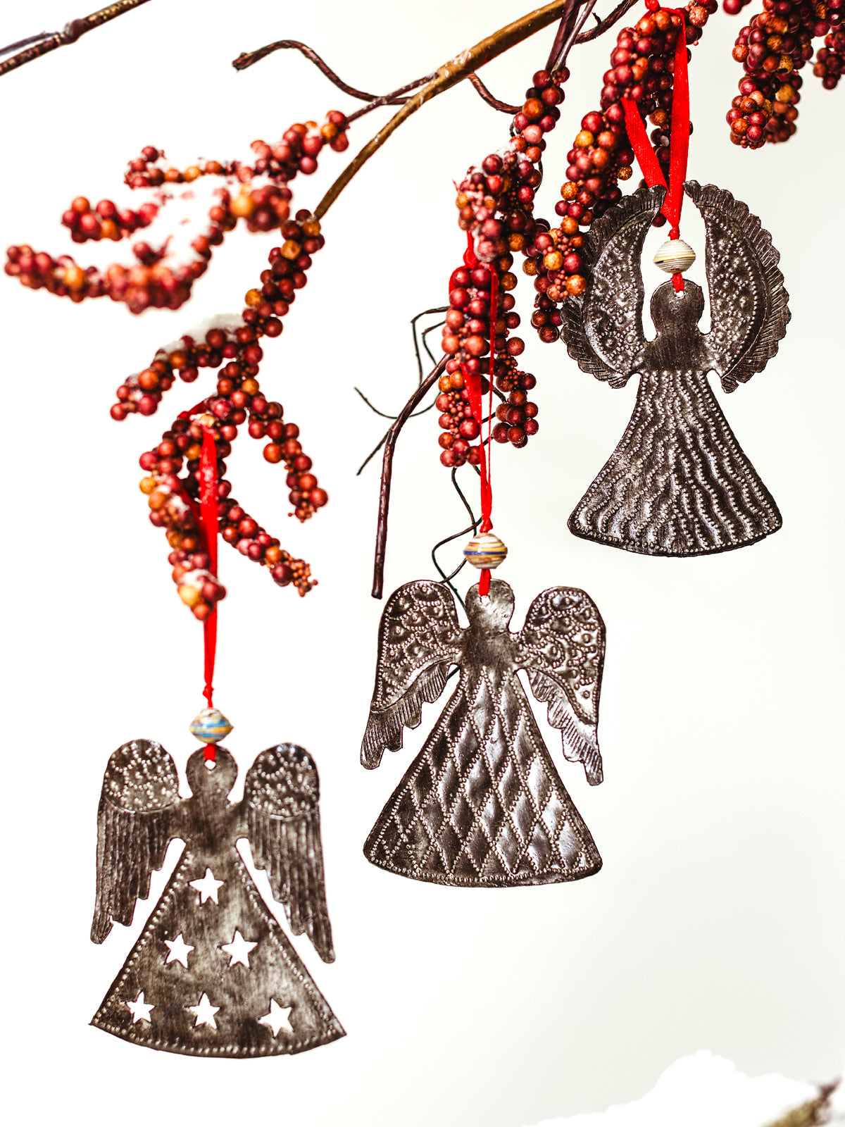 Handcrafted metal angel ornaments hanging from a branch decorated with red berries, featuring intricate patterns and red ribbon hangers, perfect for holiday decor.