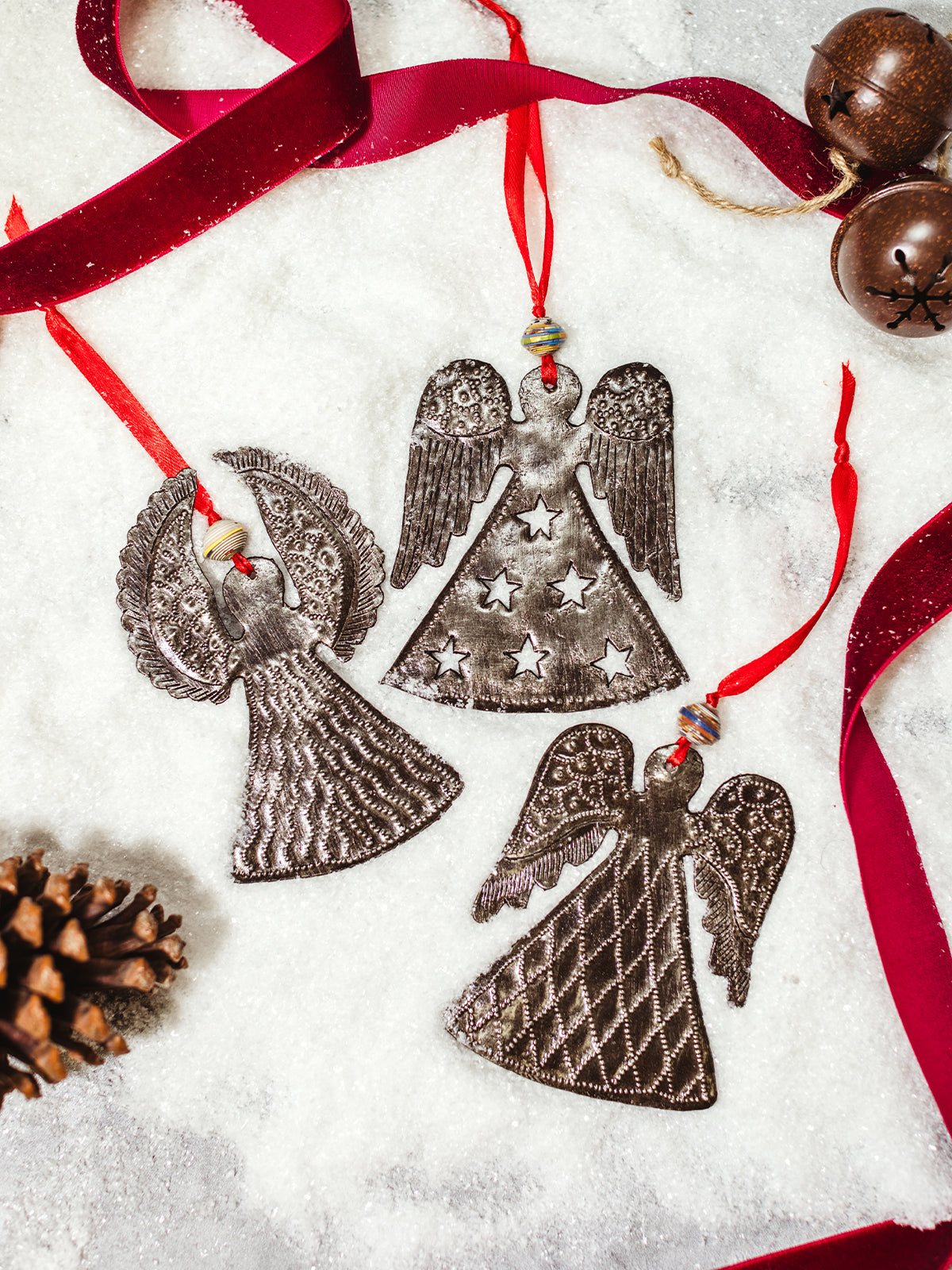 Three handmade metal angel ornaments with unique designs, lying on artificial snow, adorned with red ribbon hangers and paper beads for a festive touch.