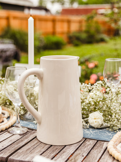Off white ceramic pitcher placed on outdoor dinning table in backyard setting.