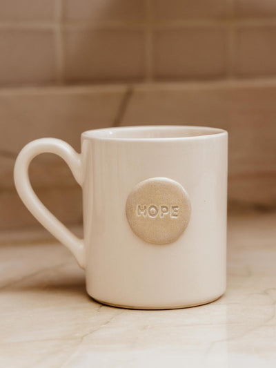 Ceramic white mug with circular ceramic piece that says "hope" sitting on white counter top.