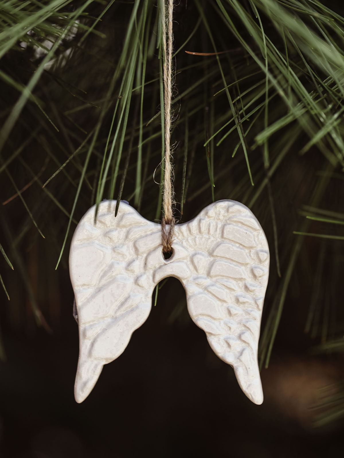 Handmade ceramic angel wings ornament with textured detail, hanging on a pine tree branch