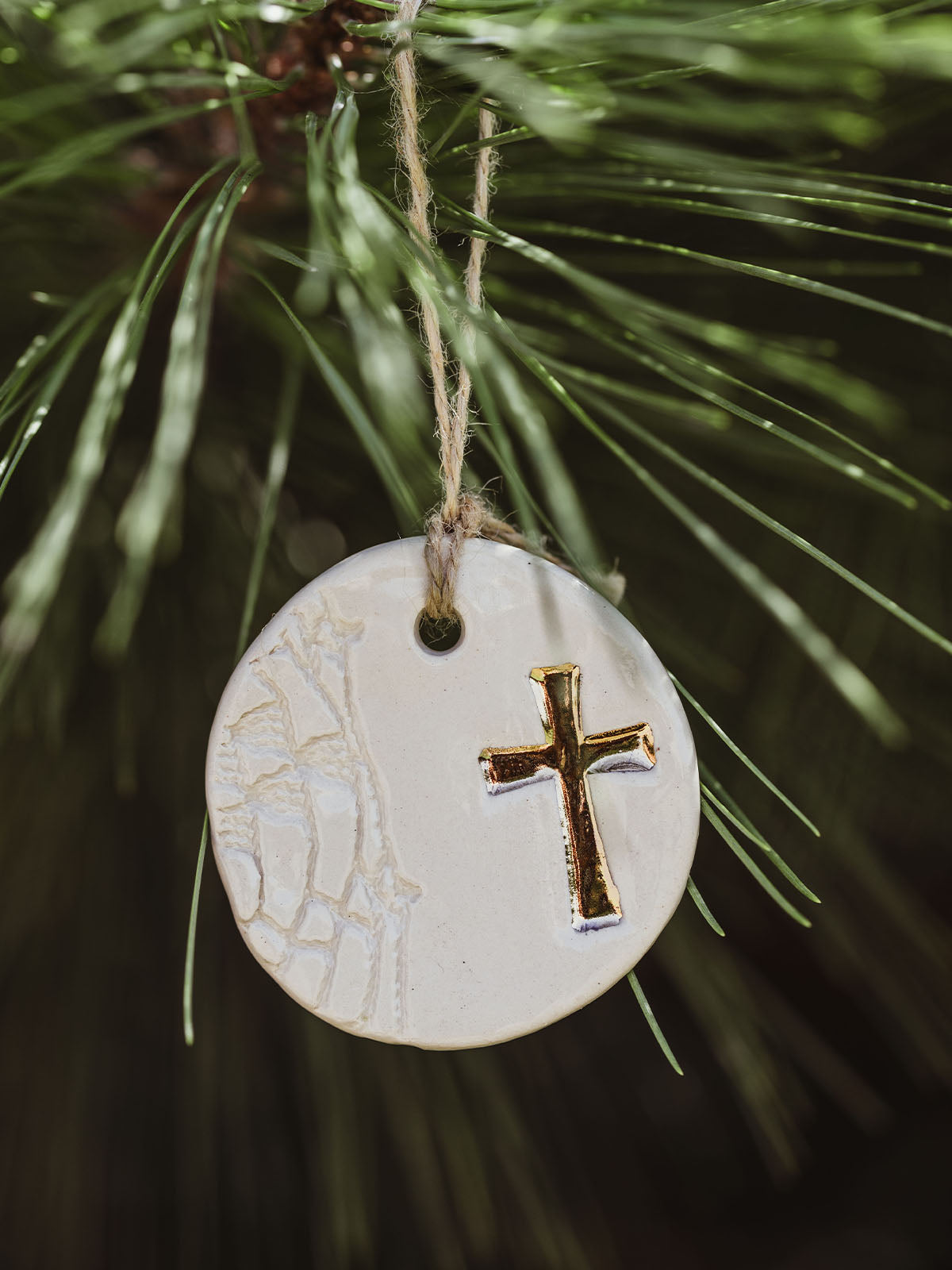 Round ceramic ornament featuring a gold cross design, hanging from a pine tree branch with natural twine.