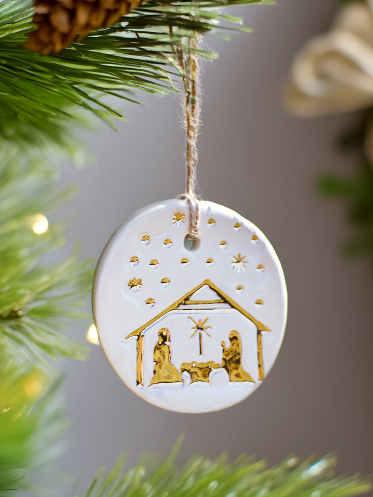 Handmade ceramic Christmas ornament depicting a nativity scene with gold accents, hanging on a green pine tree.