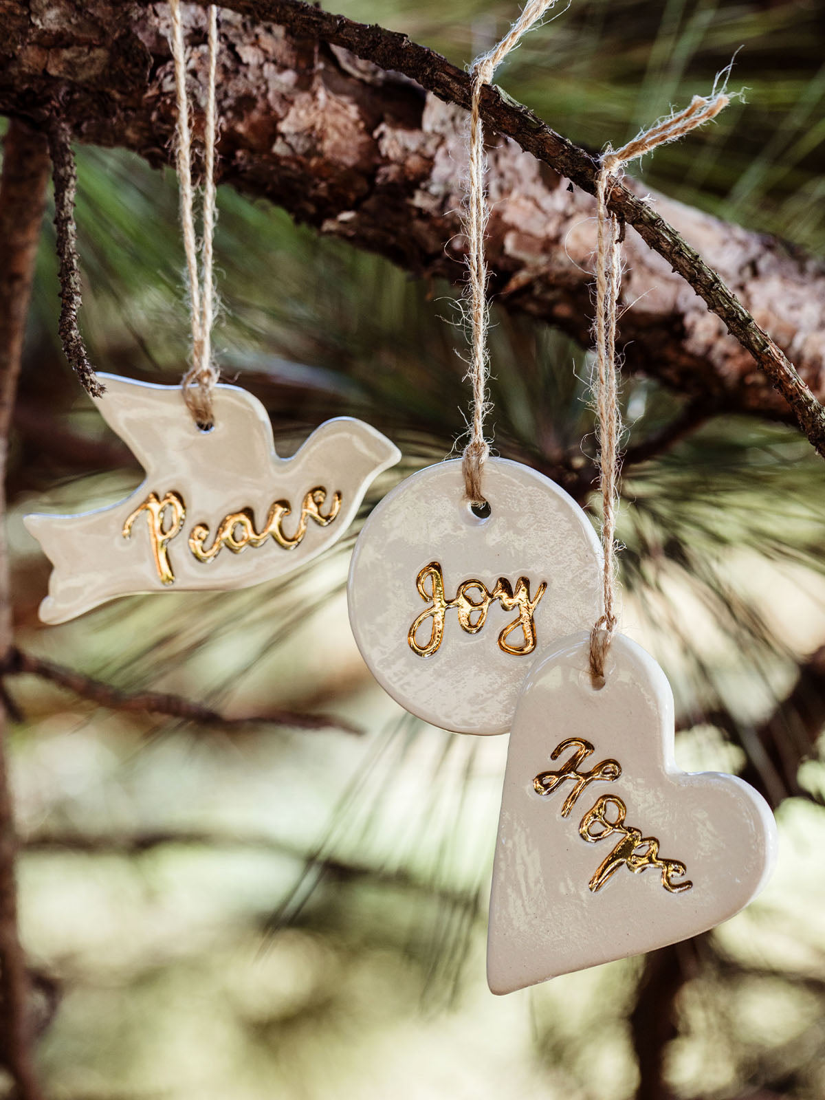 Handcrafted ceramic Christmas ornaments hanging from a tree branch, featuring a white dove with the word 'Peace,' a round ornament with 'Joy,' and a heart-shaped ornament with 'Hope,' all with gold accents. Made by Prodigal Pottery at Joffa.com.