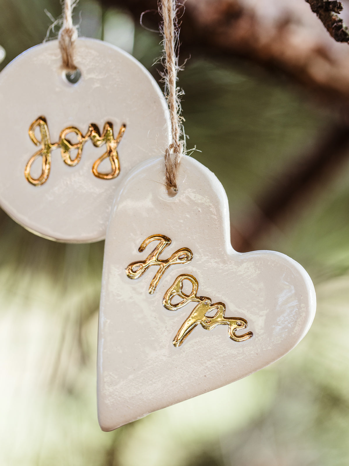 Close-up of handcrafted white ceramic Christmas ornaments with the words 'Hope' on a heart and 'Joy' on a round piece, featuring gold detailing. Ethically made by artisans at Prodigal Pottery, available at Joffa.com.