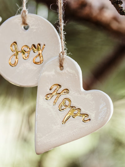 Close-up of handcrafted white ceramic Christmas ornaments with the words 'Hope' on a heart and 'Joy' on a round piece, featuring gold detailing. Ethically made by artisans at Prodigal Pottery, available at Joffa.com.