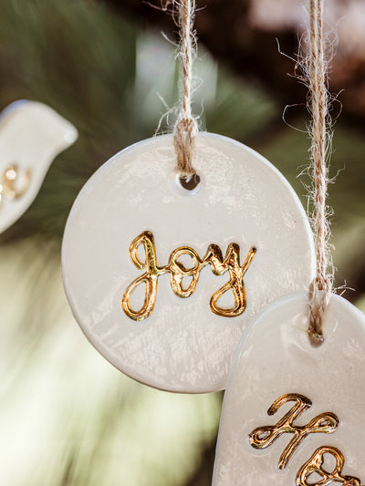 Detailed view of a handcrafted round ceramic ornament featuring the word 'Joy' in gold script, hanging from a twine string. Ethically crafted by Prodigal Pottery, sold at Joffa.com.