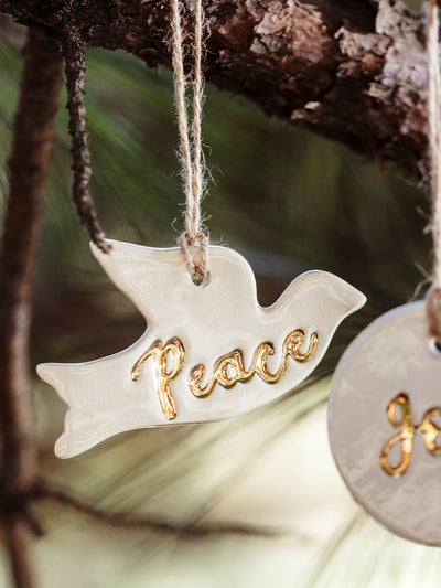 Handmade white ceramic dove ornament with the word 'Peace' in gold accents, hanging from a tree branch. A unique holiday decoration by Prodigal Pottery, available at Joffa.com.