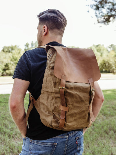 Man wearing a tan waxed canvas backpack with leather straps, standing outdoors. Durable and stylish backpack for outdoor adventures and everyday carry.