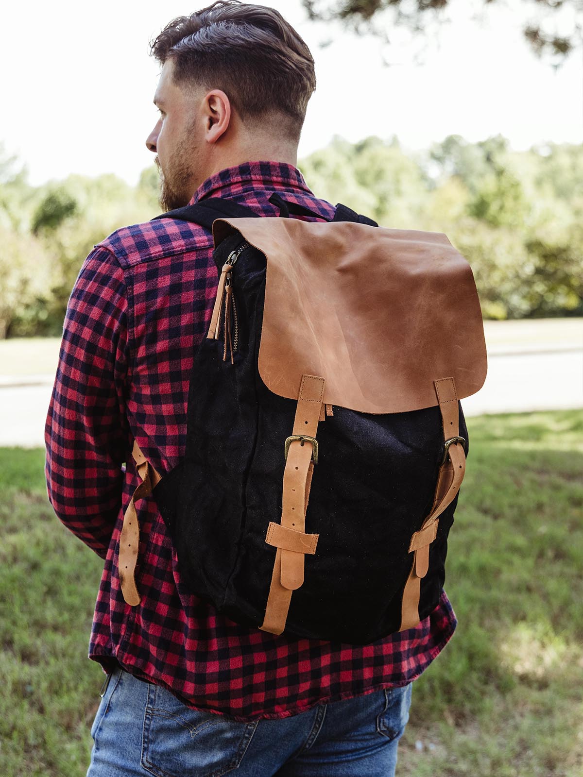 Man in a plaid shirt wearing a black canvas backpack with leather straps. Classic and rugged backpack design for casual or outdoor adventures.