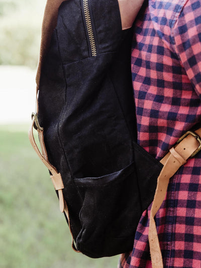 Side view of a black canvas backpack with leather detailing, worn by a person in a plaid shirt. Functional and fashionable backpack for all occasions.