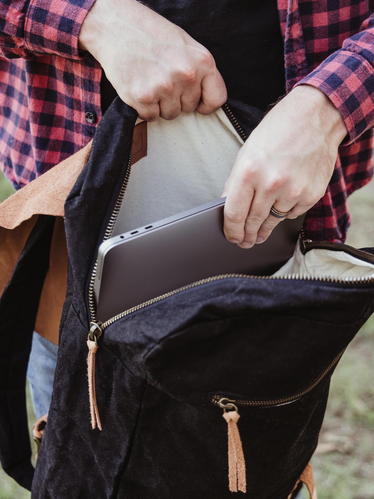 Person in a plaid shirt placing a laptop into a black waxed canvas backpack. Functional and spacious laptop bag for work or school.