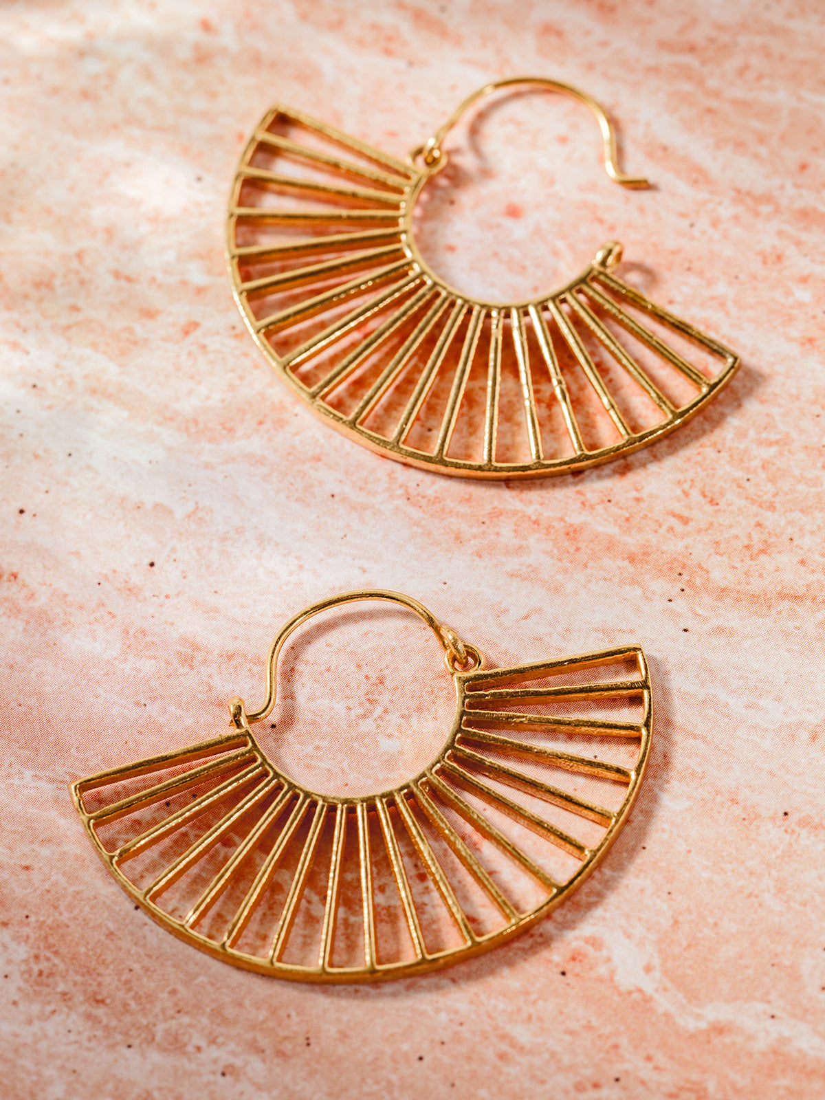 Golden fan-shaped hoop earrings with delicate openwork design, displayed on a warm peach-toned textured background, perfect for bohemian and elegant styles.