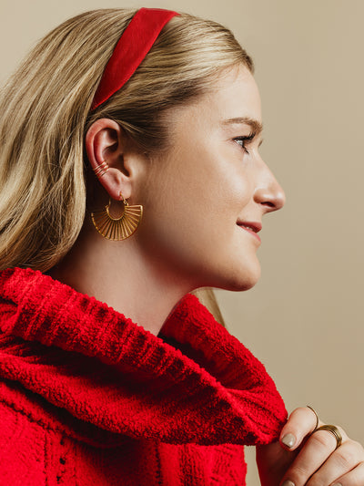 Side profile of a woman wearing gold fan-shaped earrings and stackable gold rings, styled with a bold red sweater and matching red headband, highlighting festive and handcrafted artisan jewelry for the holiday season.