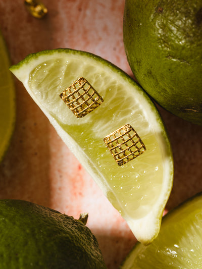 Gold square stud earrings with an intricate lattice design displayed on a fresh lime slice, showcasing their textured and elegant craftsmanship.