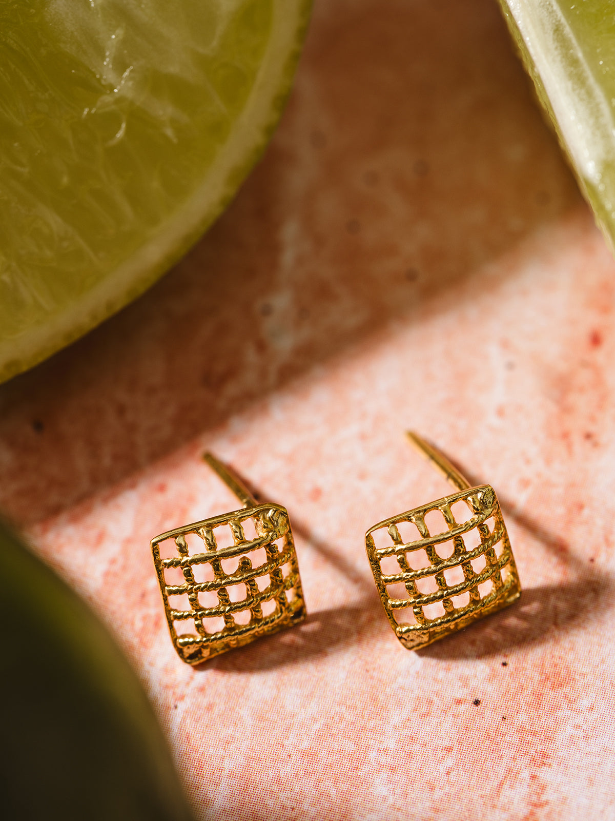 Close-up of gold square stud earrings with a textured lattice pattern, placed on a warm-toned background, perfect for a chic and minimalist style.