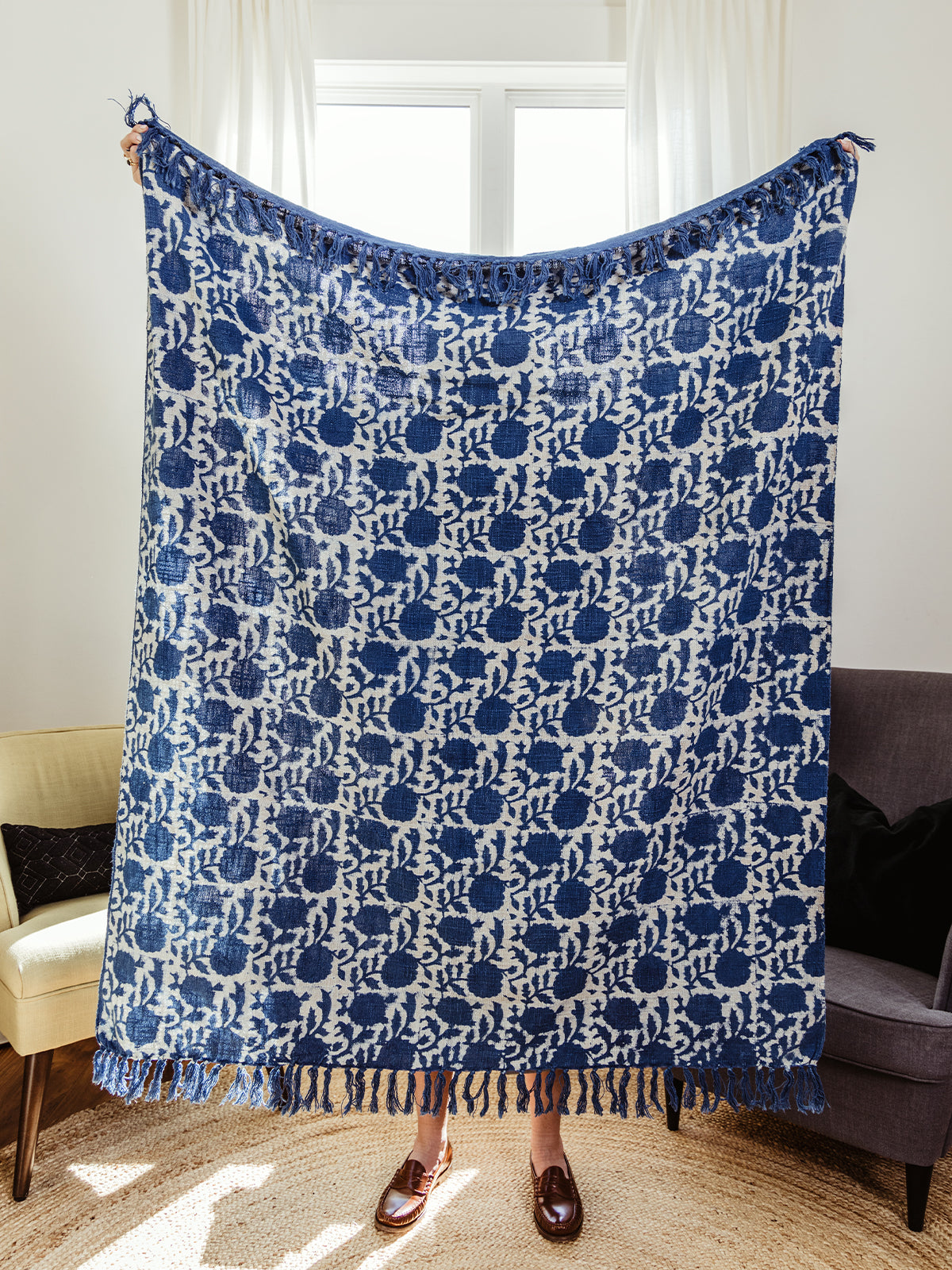 Person holding up a full-size handwoven blue and cream throw blanket with floral patterns and tassel fringe. Ethically made and perfect for adding texture and warmth to any home.