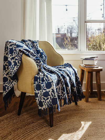 Stylish living room featuring a beige armchair with a handwoven blue and cream floral throw blanket. Natural light streams through the window, highlighting the cozy ambiance.