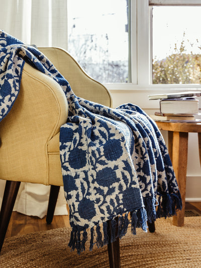 Handwoven blue and cream floral throw blanket draped over a beige accent chair in a sunlit living room. The cozy texture and intricate pattern add warmth and elegance to the space.