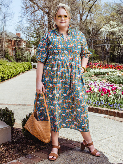 Woman wearing a vibrant floral midi dress with puff sleeves and a ruffled collar, styled with brown heeled sandals, a woven tan shoulder bag, and yellow-tinted sunglasses. She stands in a garden pathway with blooming tulips in the background.
